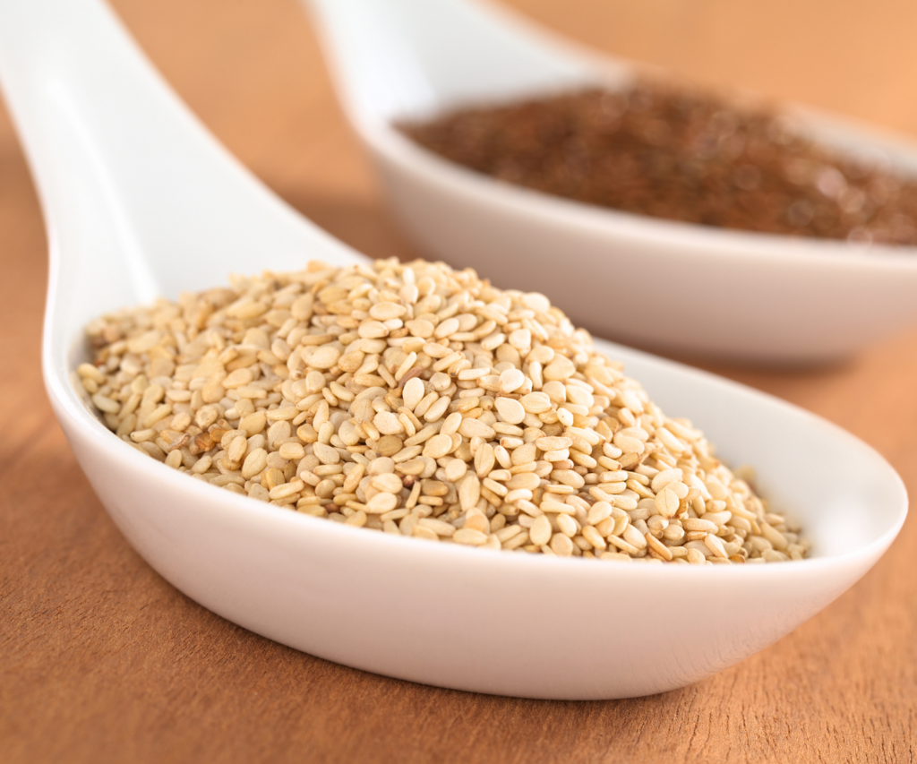 Two spoons filled with sesame seeds. One has hulled sesame seeds, one has darker sesame seeds that are not hulled. 